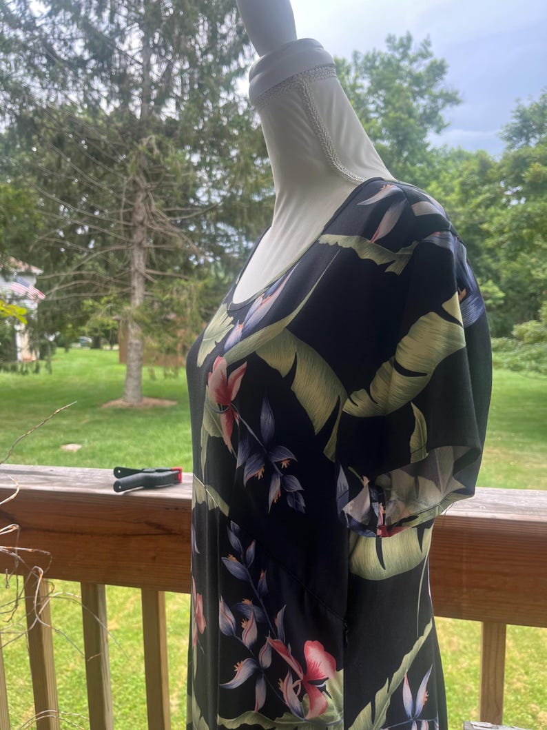 K&ouml;nnte beinhalten: Schwarzes Kleid mit tropischem Blumendruck mit rosa, blauen und gr&uuml;nen Blumen und Bl&auml;ttern. Das Kleid hat kurze &Auml;rmel und einen Rundhalsausschnitt. Das Kleid wird auf einer Schaufensterpuppe pr&auml;sentiert.