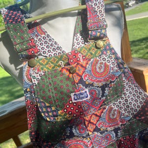 May include: Close-up of colorful patchwork overalls with paisley and floral patterns. The overalls feature adjustable straps, brass-colored buttons, and a front pocket with a "BIG SMITH" label. The fabric is a mix of red, green, blue, and black.