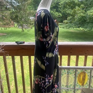 K&ouml;nnte beinhalten: Schwarzes Maxikleid mit tropischem Blumendruck mit rosa, lila und gr&uuml;nen Blumen und Bl&auml;ttern. Das Kleid hat kurze &Auml;rmel und eine flie&szlig;ende Silhouette. Das Kleid wird auf einer Schaufensterpuppe pr&auml;sentiert.