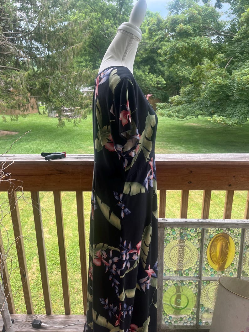 K&ouml;nnte beinhalten: Schwarzes Maxikleid mit tropischem Blumendruck mit rosa, lila und gr&uuml;nen Blumen und Bl&auml;ttern. Das Kleid hat kurze &Auml;rmel und eine flie&szlig;ende Silhouette. Das Kleid wird auf einer Schaufensterpuppe pr&auml;sentiert.