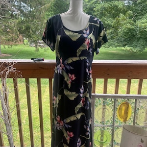 K&ouml;nnte beinhalten: Ein schwarzes Maxikleid mit tropischem Blumendruck, mit rosa Bl&uuml;ten und gr&uuml;nen Bl&auml;ttern. Das Kleid hat kurze &Auml;rmel und einen Rundhalsausschnitt. Der Stoff f&auml;llt bis zum Boden und erzeugt eine flie&szlig;ende Silhouette. Das Kleid wird auf einer Schaufensterpuppe pr&auml;sentiert.