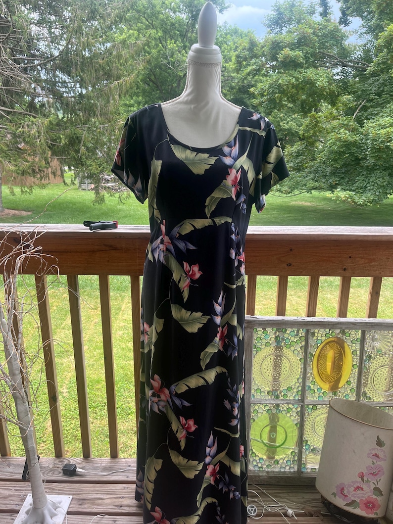 K&ouml;nnte beinhalten: Ein schwarzes Maxikleid mit tropischem Blumendruck, mit rosa Bl&uuml;ten und gr&uuml;nen Bl&auml;ttern. Das Kleid hat kurze &Auml;rmel und einen Rundhalsausschnitt. Der Stoff f&auml;llt bis zum Boden und erzeugt eine flie&szlig;ende Silhouette. Das Kleid wird auf einer Schaufensterpuppe pr&auml;sentiert.