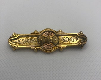 May include: A gold-toned brooch with an intricate design. The center of the brooch features a circular design with a raised, textured pattern. The brooch is in the shape of a rectangle with rounded corners.