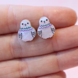 May include: A pair of stud earrings shaped like small birds. Each earring is made of a translucent, glittery white material with hand-drawn black outlines for the wings and eyes, and small purple and green decorative lines across the chest area.
