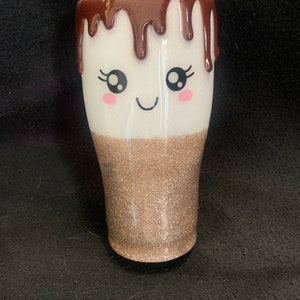 May include: A white and gold glitter tumbler with a brown chocolate-like design dripping down the side. The tumbler has a cute cartoon face with eyelashes and a smile.