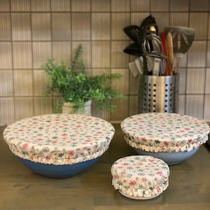 May include: Three reusable bowl covers with a snowflake pattern in shades of red, blue, and green on a cream background. The covers are made of fabric and have a ruffled edge. The covers are sitting on a countertop.
