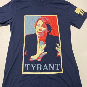 May include: A navy blue t-shirt with a graphic of a woman in a red, white, and blue color scheme. The graphic is in a square frame with the word "TYRANT" printed below it. The t-shirt has a small American flag patch on the right sleeve.