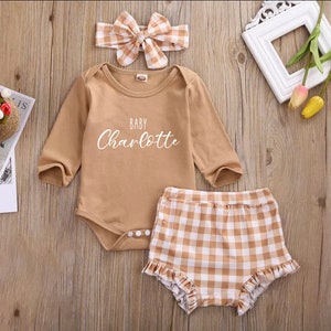 May include: A three-piece baby outfit in brown and white. The outfit includes a long-sleeved bodysuit with the text "BABY Charlotte", a headband with a bow, and checkered shorts with ruffles.