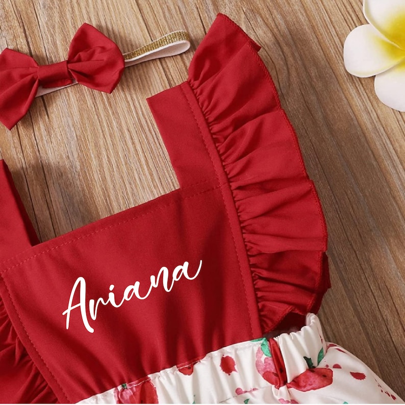 May include: Red and white baby onesie with a ruffled red bodice and a white bow headband. The onesie has a white floral print with red strawberries and the name "Ariana" is embroidered on the bodice.