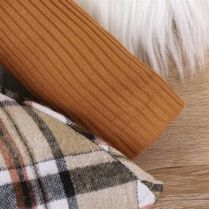 May include: Close-up of a tan ribbed knit long sleeve top with a plaid pattern. The plaid pattern features black, white, and brown colors. The top is made of a soft, comfortable material.