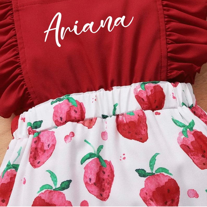 May include: A red and white baby onesie with a ruffled neckline and a strawberry print. The onesie has the name "Ariana" embroidered on the front.