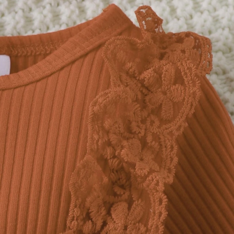 May include: Close-up of a rust-colored ribbed knit top with delicate lace detailing on the shoulder. The lace is a matching shade of rust, creating a cohesive and textured look. The top has a round neckline.