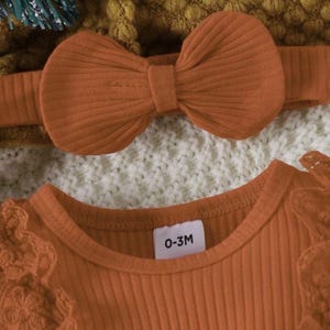 May include: An orange baby outfit set, including a ribbed top with lace sleeves and a matching bow headband. The top has a tag that reads "0-3M". The set is displayed on a textured white and yellow background.