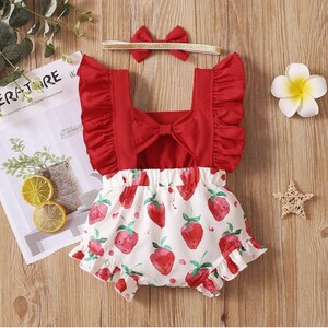 May include: Red and white baby onesie with a strawberry print and a red bow on the back. The onesie has ruffles on the shoulders and around the legs. A gold headband with a red bow is included.