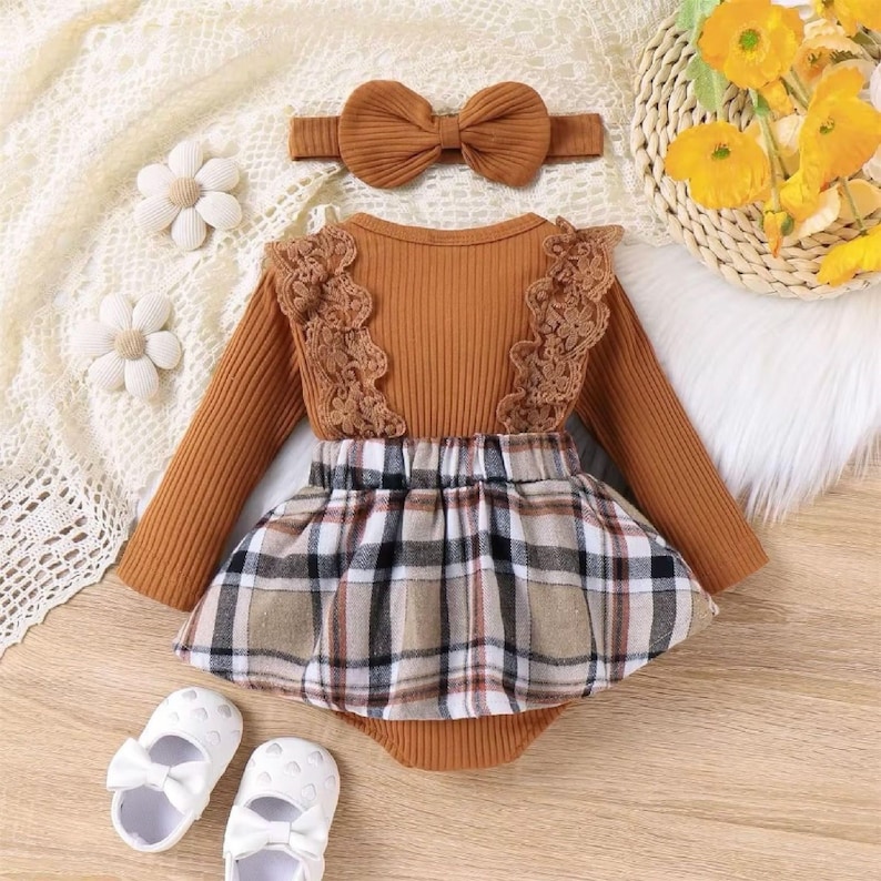 May include: A baby girl's outfit set featuring a brown long-sleeved bodysuit with lace accents, a plaid skirt, and a matching headband with a bow. White baby shoes with bows are also included.
