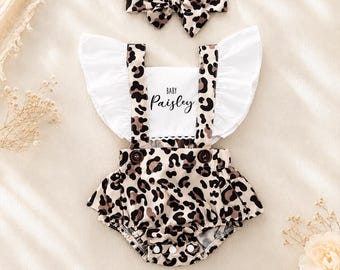 May include: A leopard print romper with white ruffled sleeves and a matching headband. The romper features a white chest panel with the text Baby Paisley printed in black, two brown buttons at the waist, and snap closures at the bottom.