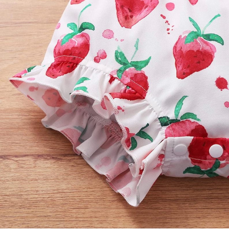 May include: White baby onesie with a ruffled hem and a strawberry print. The onesie has a button closure at the bottom.