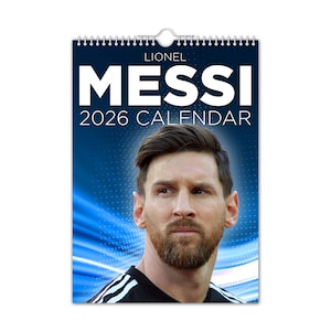 May include: A wall calendar featuring Lionel Messi with the text "MESSI 2026 CALENDAR" in large white letters. The calendar has a blue background with a portrait of Messi. The calendar is held together with a silver spiral binding.
