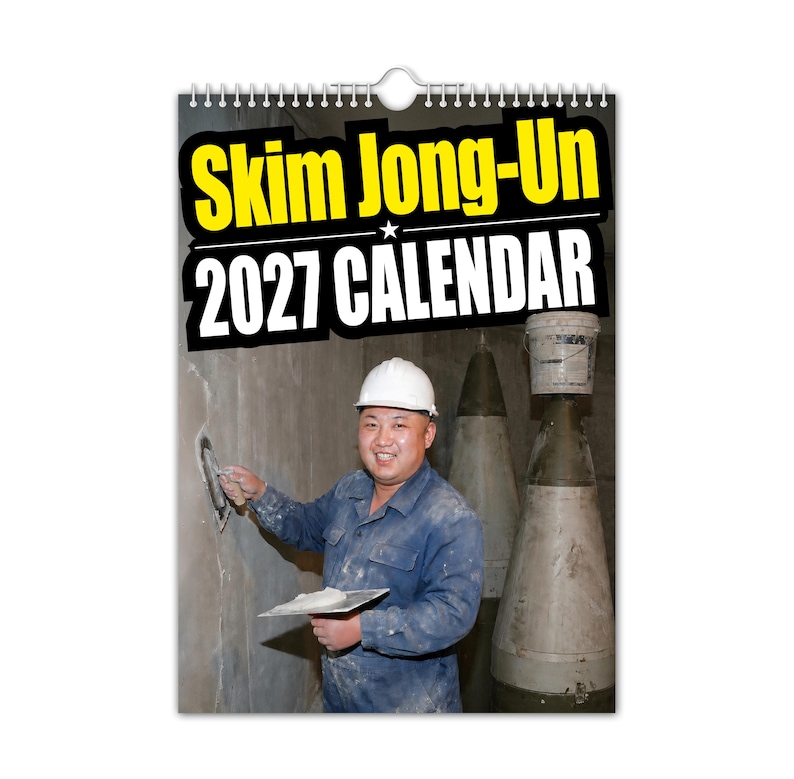 Pu&ograve; includere: Un calendario da parete con rilegatura a spirale e la scritta "Skim Jong-Un 2027 CALENDAR" in giallo e bianco. L'immagine mostra un uomo in tuta blu da lavoro e casco bianco, che applica intonaco a un muro. Due grandi oggetti cilindrici sono sullo sfondo.