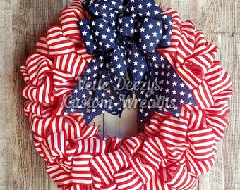 May include: A patriotic wreath featuring red and white striped ribbon, forming a circular shape. A large navy blue bow with white stars is centered at the top. The text "Vette Deezy's Custom Wreaths" is visible.