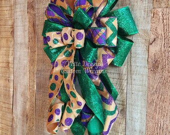 May include: A green, gold, and purple glittery bow with polka dots and chevron patterns. The bow is tied with a loop and is hanging on a wooden background. "Vette Deezy's Custom Wreaths" is written on the bow.