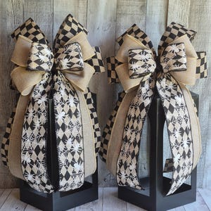May include: Two decorative bows crafted from wired ribbon featuring a black and white diamond pattern, a spider and web print, and solid tan burlap sections. Each bow is mounted atop a black metal lantern frame with an open-sided design.