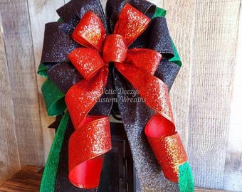 May include: A decorative bow with black, red, and green ribbons. The bow has a glittery texture and is centered with a large red bow. The text "Vette Deezy's Custom Wreaths" is visible on the bow.