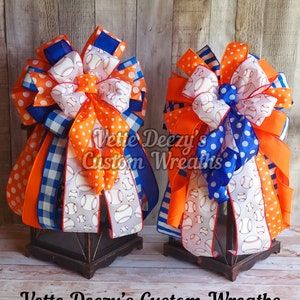 May include: Two orange, blue, and white baseball-themed bows on black lanterns. The bows are made with various ribbons, including polka dot, plaid, and baseball print. The lanterns are on a wooden surface. Vette Deezy's Custom Wreaths is written on the surface.
