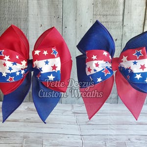 May include: Two red, white, and blue star patterned hair bows. The bows are made of fabric and have a layered design. The bows are tied together in the center with a white ribbon.