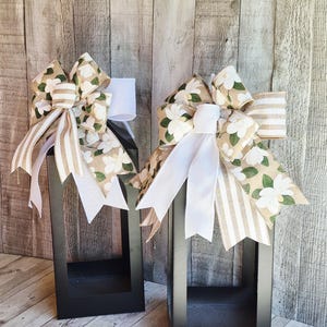 May include: Two black rectangular lanterns with decorative bows. The bows feature white magnolia flowers, tan and white striped ribbon, and solid white ribbon. The lanterns are empty and sit on a light wood surface.