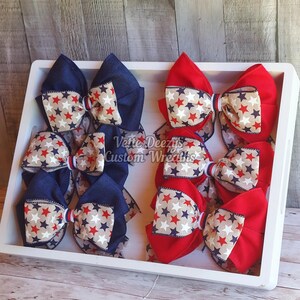 May include: Six handmade hair bows in red and blue denim fabric with white and red stars. The bows are arranged in a white tray. The text "Vette Deezy's Custom Wreaths" is visible in the center of the image.