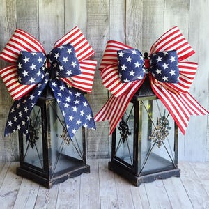 May include: Two black metal lanterns with glass panels, adorned with patriotic bows. The bows feature a blue center with white stars and red and white striped ribbons, creating a festive American flag theme.