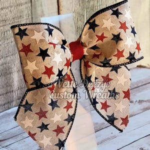 May include: A large bow made from burlap ribbon with red, white, and blue stars. The bow is tied with a red ribbon in the center.
