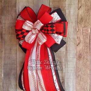 May include: A red, white, and black baseball-themed bow with a white ribbon with red stitching and a black ribbon with white baseballs. The bow is tied with a black ribbon and has the text "Vette Deezy's Custom Wreaths" on it.