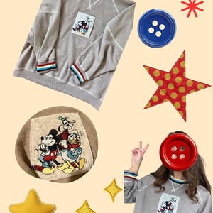May include: A beige long-sleeve sweatshirt featuring a rectangular patch with embroidered Mickey Mouse, Goofy, and Donald Duck characters. The collar and cuffs have striped red, white, blue, and yellow trim. Includes decorative star and button shapes.