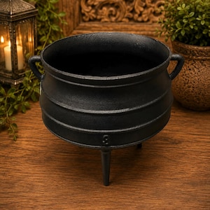 May include: A black cast iron cauldron with a wide circular opening, a rounded body featuring three horizontal ridges, two small side handles, and three tapered legs extending from the base. The surface has a matte, textured finish.
