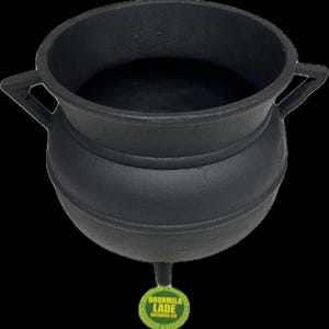 May include: Black cast iron cauldron with two handles. The cauldron has a rounded bottom and a wide opening. The cauldron is sitting on a white surface. The text "ORUNMILA LADE BOTANICA LLC" is printed on a green label attached to the cauldron.