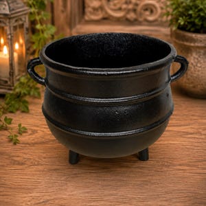 May include: A black cast iron cauldron featuring a rounded body with two side handles and three short legs. The exterior surface has a textured finish with two horizontal ridges encircling the middle section of the pot, creating a classic design.