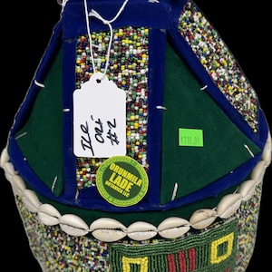May include: A handcrafted, beaded hat with a unique design. The hat features a combination of green, blue, and multicolored beads, with a row of white shells along the base. A tag is attached to the hat.