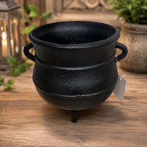 May include: A black cast iron cauldron with a rounded body, a wide open top, and three short legs. The pot features two small side handles and a textured surface finish. A small white rectangular paper tag is attached to one of the side handles.