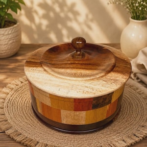 May include: A round wooden container with a lid featuring a central knob. The exterior body is constructed from a mosaic of rectangular wood pieces in varying shades of brown, tan, orange, and dark red, while the lid displays a smooth wood grain.