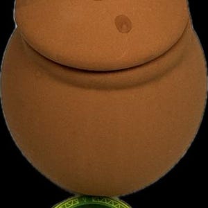 May include: A small, terracotta-colored jar with a lid. The jar has a round body and a slightly flared opening. The lid has a small knob on top. The jar is labeled "Orunmila Lade Botanica LLC".