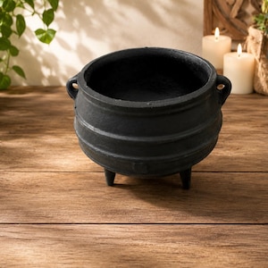 May include: A matte black cast iron cauldron with a rounded body, three short legs, and two small side handles. The pot features a wide circular opening and a textured surface, presenting a deep, hollow interior and a sturdy, heavy-duty construction.