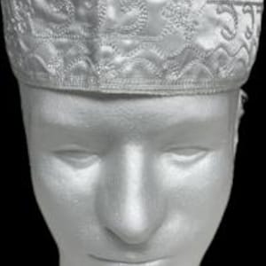 May include: A white embroidered kufi hat with intricate patterns. The hat is made of a shiny fabric and features detailed stitching along the edges and top. The hat is displayed on a white mannequin head against a black background.