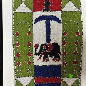May include: A beaded wall hanging with a geometric design featuring a red face, a blue and white symbol, and a black elephant. The background is green and white.