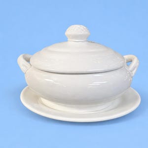 May include: White ceramic soup tureen with a lid and two handles, resting on a matching saucer. The lid has a decorative knob. The set is all white and sits against a light blue background.