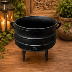 May include: A black cast iron cauldron with a rounded body, three tapered legs, and two small side handles. The exterior features a raised number 2 embossed on the front surface. The vessel has a wide, open top and a smooth, dark finish.