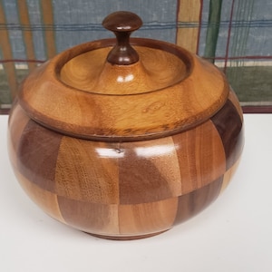 May include: A wooden bowl with a lid, made of different types of wood. The bowl is round and has a checkered pattern. The lid has a small knob on top.