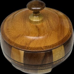 May include: A round wooden container with a lid featuring a small knob handle. The body of the container is constructed from alternating vertical segments of light and dark brown wood, showcasing a polished finish and natural wood grain patterns.