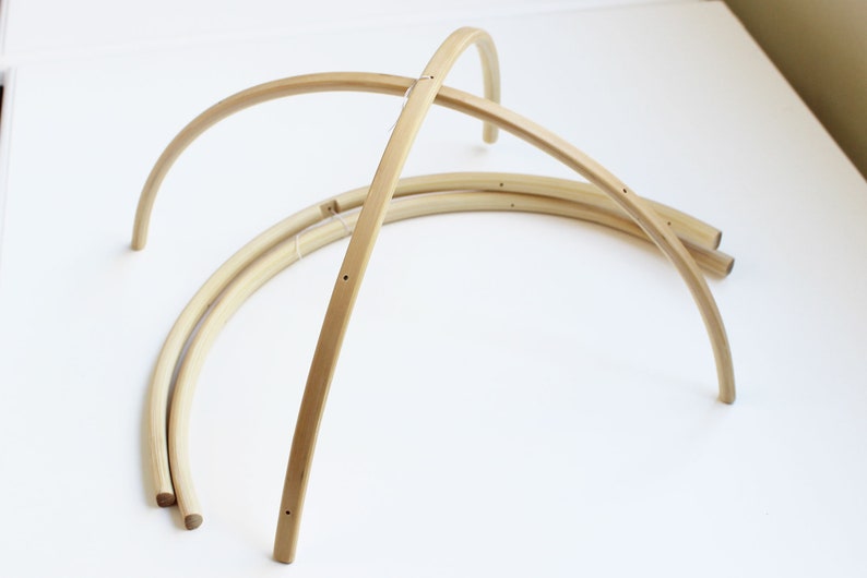May include: Three unfinished wooden hoops, each with a slight curve, are stacked on top of each other. The hoops are made of light brown wood and have small holes drilled into them.
